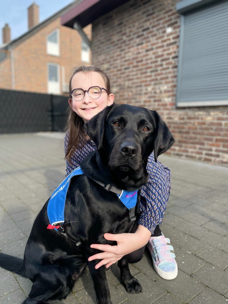 Whisper, een zwarte labrador, zit voor Dina, een lachend meisje, die haar arm rond de hond heeft liggen.